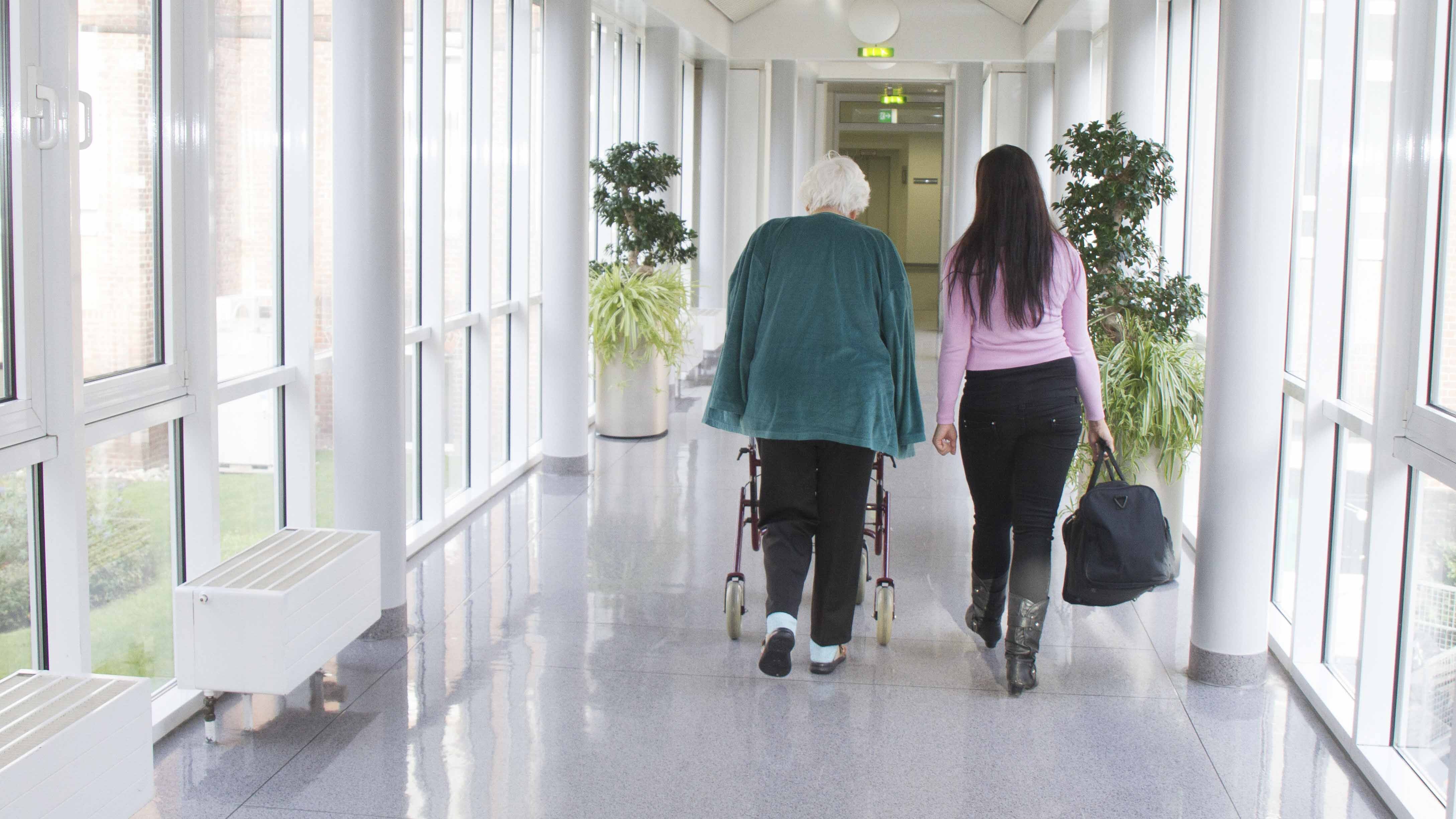 Auf dem Bild ist ein Flur des Krankenhauses zu sehen, auf dem mittig zwei Personen mit dem Rücken zur Kamera lang laufen. Eine ältere Dame (li.) läuft am Rolator und wird von einer jüngeren Dame (re.) begleitet. 