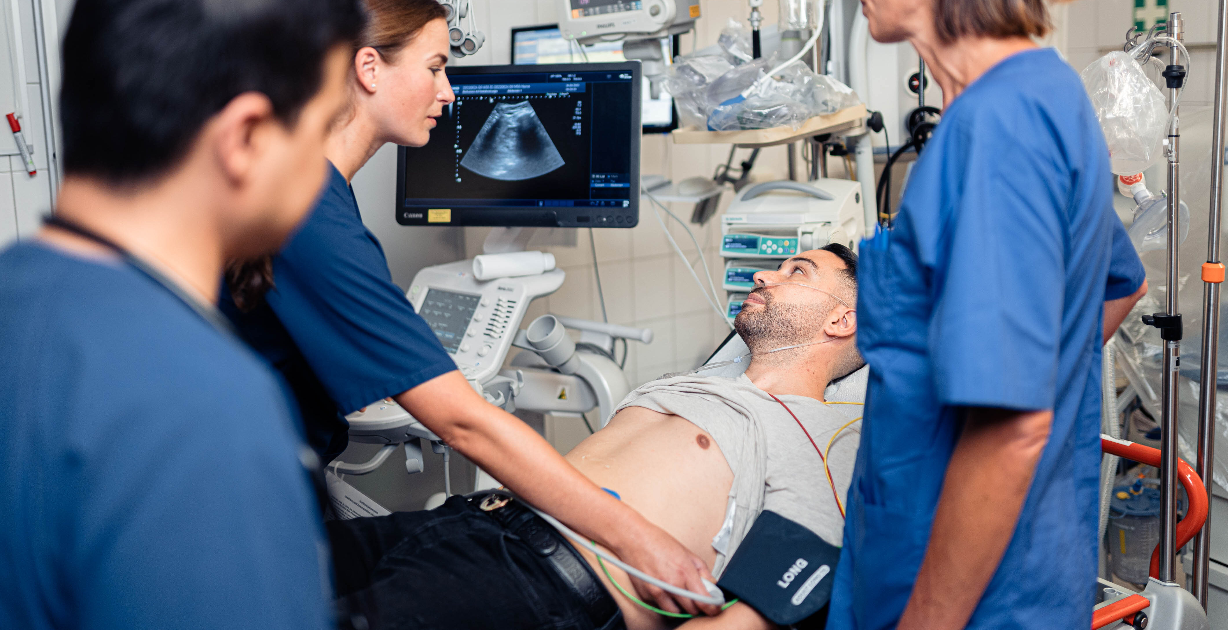 Auf dem Bild ist der Schockraum der Notaufnahme abgebildet. Ein Patient liegt in der Mitte auf der Liege, um ihn herum stehen Notfallsanitäter, ein Arzt und ein Pfleger. Außerdem ist medizinische Ausrüstung und Material zu sehen. 